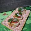Vue en biais d’une assiette rectangulaire à motif floral rouge, garnie de trois canapés sur pain croustillant. Chaque bouchée associe tomate fraîche, sardine, pointe de piment et persil, dans une harmonie simple et savoureuse. En arrière-plan, un set de table vert aux motifs tropicaux de feuilles et d’oiseaux apporte une touche exotique et conviviale à l’ensemble.
