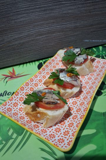 Vue en biais d’une assiette rectangulaire à motif floral rouge, garnie de trois canapés sur pain croustillant. Chaque bouchée associe tomate fraîche, sardine, pointe de piment et persil, dans une harmonie simple et savoureuse. En arrière-plan, un set de table vert aux motifs tropicaux de feuilles et d’oiseaux apporte une touche exotique et conviviale à l’ensemble.