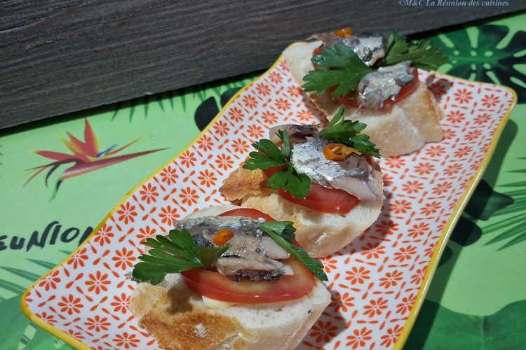 Vue en biais d’une assiette rectangulaire à motif floral rouge, garnie de trois canapés sur pain croustillant. Chaque bouchée associe tomate fraîche, sardine, pointe de piment et persil, dans une harmonie simple et savoureuse. En arrière-plan, un set de table vert aux motifs tropicaux de feuilles et d’oiseaux apporte une touche exotique et conviviale à l’ensemble.