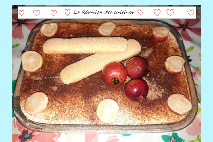 Dessert créatif façon tiramisu, présenté dans un plat rectangulaire en verre. Le dessus est saupoudré de cacao et décoré avec des fruits goyavier et deux biscuits cuillère croisés. Le plat repose sur une nappe fleurie. En arrière-plan, une bannière "La Réunion des cuisines" encadrée de cœurs rouges, avec une illustration tropicale mêlant palmier et drapeau italien en forme de cœur. Ambiance festive et métissée, célébrant la fusion culinaire entre La Réunion et l’Italie