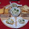 Plateau apéritif sur une nappe rouge décorée de motifs dorés et blancs en forme d’étoiles et de flocons. Sur une planche ronde en bois, deux verrines carrées remplies de chips, un petit bol blanc contenant des graines de jacques pilées et garnies d’herbes, ainsi qu’une coupelle avec une préparation à base d’oignons. Devant, deux chips garnies sont posées sur des serviettes décoratives. L’ensemble évoque une ambiance de fête conviviale et gourmande
