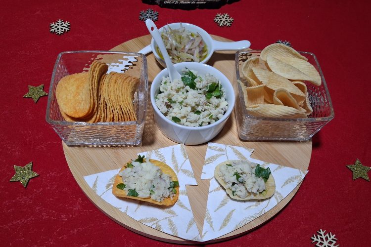 Plateau apéritif sur une nappe rouge décorée de motifs dorés et blancs en forme d’étoiles et de flocons. Sur une planche ronde en bois, deux verrines carrées remplies de chips, un petit bol blanc contenant des graines de jacques pilées et garnies d’herbes, ainsi qu’une coupelle avec une préparation à base d’oignons. Devant, deux chips garnies sont posées sur des serviettes décoratives. L’ensemble évoque une ambiance de fête conviviale et gourmande