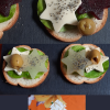 Trois bouchées festives présentées sur des tranches de pain, décorées de feuilles vertes, étoiles de fromage blanc et de betterave, olives vertes et graines de pavot. Chaque composition évoque une ambiance de fête, avec une touche créative et végétale. En bas, une bouchée repose sur une serviette en forme de sapin, renforçant l’esprit de Noël.