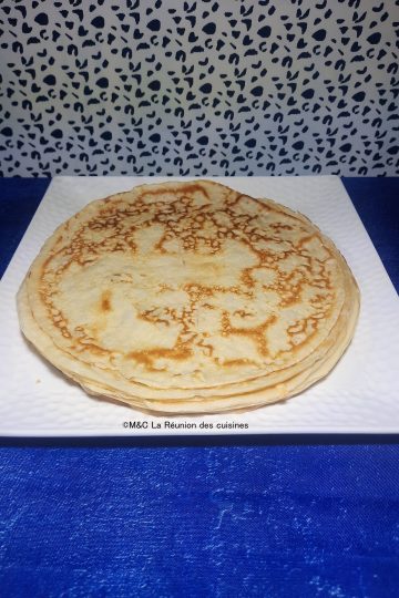Une pile de crêpes fines et dorées, présentées sur une assiette carrée blanche, posée sur une nappe bleue éclatante. Les crêpes montrent une cuisson parfaite avec des motifs marbrés appétissants.