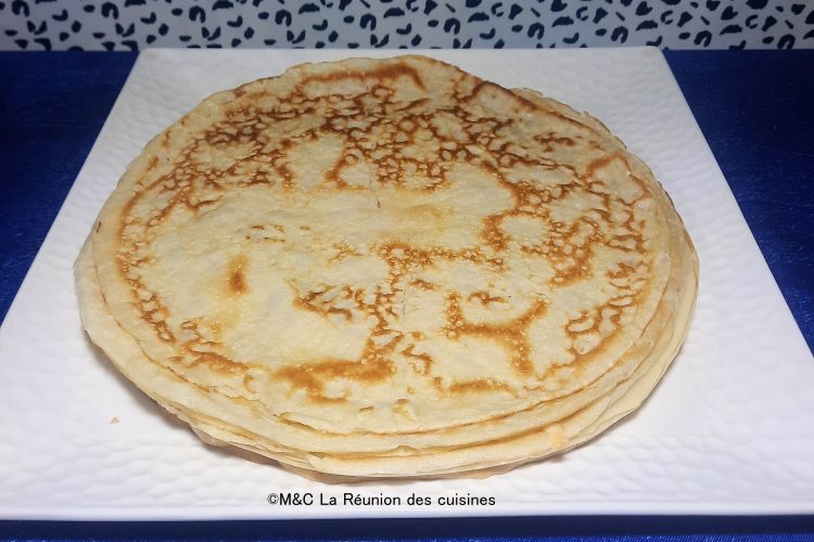 Une pile de crêpes fines et dorées, présentées sur une assiette carrée blanche, posée sur une nappe bleue éclatante. Les crêpes montrent une cuisson parfaite avec des motifs marbrés appétissants.