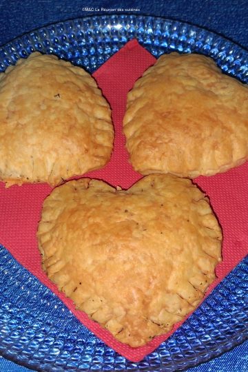Trois cœurs feuilletés dorés posés sur une serviette rouge, présentés dans une assiette en verre, sur une nappe bleue.