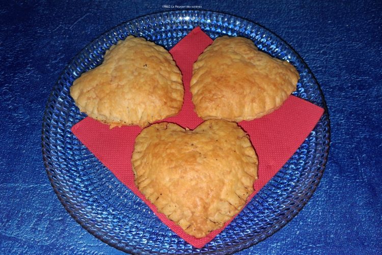 Trois cœurs feuilletés dorés posés sur une serviette rouge, présentés dans une assiette en verre, sur une nappe bleue.