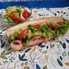 Un maxi hot-dog généreux présenté sur une serviette de table en papier fleurie, garni de tranches de jambon, de feuilles de salade verte croquante, de rondelles de tomates cerises et d'un piment rouge. À côté, un petit bol en verre contient une salade d'accompagnement fraîche avec tomates et piment.