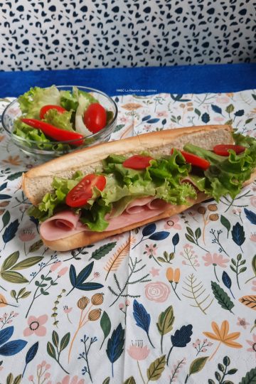 Un maxi hot-dog généreux présenté sur une serviette de table en papier fleurie, garni de tranches de jambon, de feuilles de salade verte croquante, de rondelles de tomates cerises et d'un piment rouge. À côté, un petit bol en verre contient une salade d'accompagnement fraîche avec tomates et piment.
