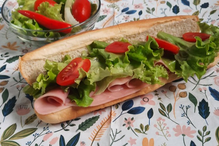 Un maxi hot-dog généreux présenté sur une serviette de table en papier fleurie, garni de tranches de jambon, de feuilles de salade verte croquante, de rondelles de tomates cerises et d'un piment rouge. À côté, un petit bol en verre contient une salade d'accompagnement fraîche avec tomates et piment.