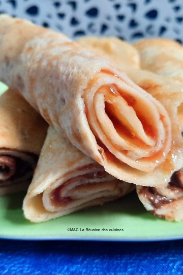 Assiette verte de crêpes express garnies de confiture et chocolat, posée sur une nappe bleue avec un fond à motifs noirs. Recette La Réunion des Cuisines.