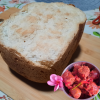 Un gros pain à la patate douce blanche entier, cuit en machine à pain (MAP), posé sur une planche en bambou sur une nappe à fleurs. En bas à droite, un ramequin de rougail saucisse créole décoré de deux fleurs de frangipanier roses. En haut à droite, une petite tasse blanche avec un motif rouge est positionnée sur la manique/le sous-plat