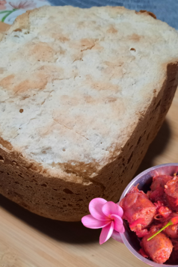 Un gros pain à la patate douce blanche entier, cuit en machine à pain (MAP), posé sur une planche en bambou sur une nappe à fleurs. En bas à droite, un ramequin de rougail saucisse créole décoré de deux fleurs de frangipanier roses. En haut à droite, une petite tasse blanche avec un motif rouge est positionnée sur la manique/le sous-plat