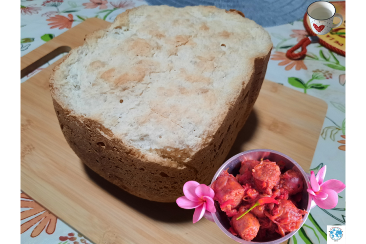 Un gros pain à la patate douce blanche entier, cuit en machine à pain (MAP), posé sur une planche en bambou sur une nappe à fleurs. En bas à droite, un ramequin de rougail saucisse créole décoré de deux fleurs de frangipanier roses. En haut à droite, une petite tasse blanche avec un motif rouge est positionnée sur la manique/le sous-plat