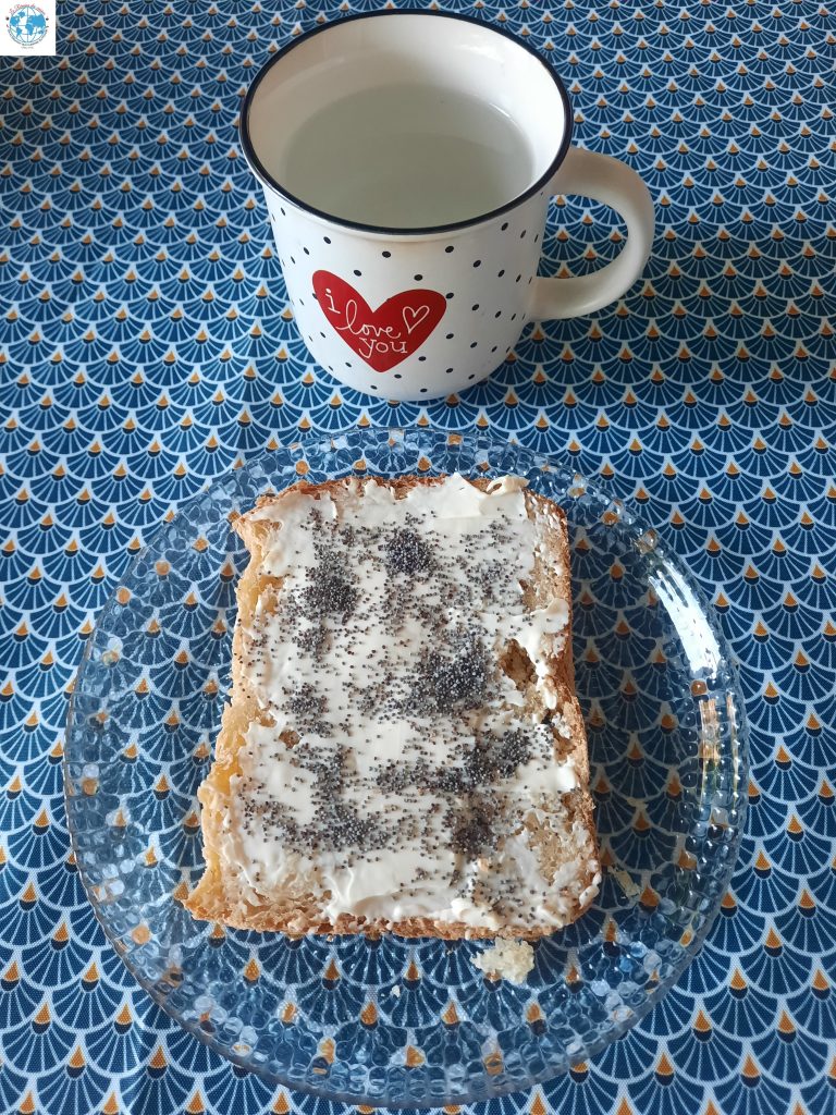 Une vue de dessus d'une tranche de pain maison à la patate douce sur une assiette en verre texturé. La tranche est tartinée de fromage à la crème et parsemée de graines de pavot. En arrière-plan, une tasse blanche à pois avec un cœur rouge est posée sur une nappe à motif géométrique bleu.