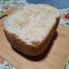Vue de dessus d'un pain à la patate douce cuit en machine à pain, posé sur une planche à découper en bois. La croûte supérieure est claire avec des nuances dorées et présente une texture légèrement craquelée et rustique.