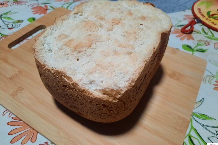 Vue de dessus d'un pain à la patate douce cuit en machine à pain, posé sur une planche à découper en bois. La croûte supérieure est claire avec des nuances dorées et présente une texture légèrement craquelée et rustique.
