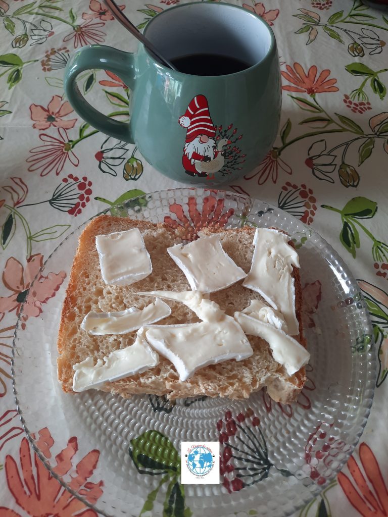 Une vue de dessus d'une tranche de pain maison à la patate douce sur une assiette en verre texturé. La tranche est généreusement garnie de morceaux de fromage crémeux, type Caprice des Dieux. En arrière-plan, une tasse de café verte ornée d'un dessin de gnome est posée sur une nappe fleurie