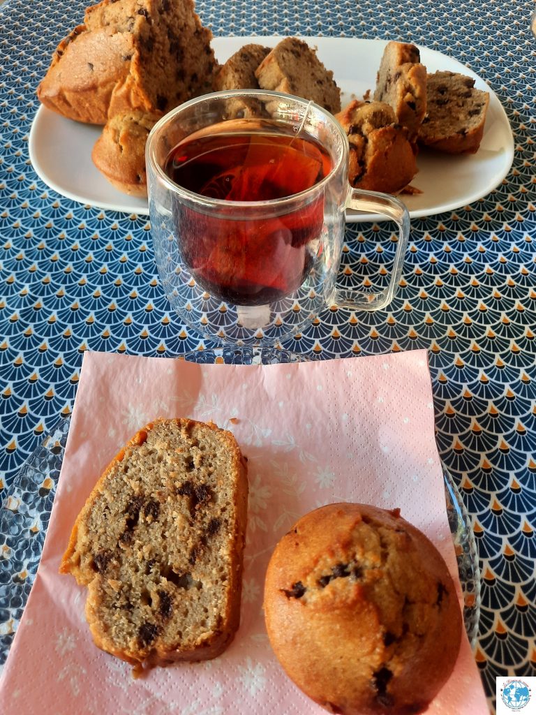 Pause gourmande avec une tranche de Cake-Dôme banane pépites et un dôme moelleux sur une serviette rose, accompagnés d'une tasse de thé chaud. Le tout posé sur une nappe bleue à motifs graphiques.