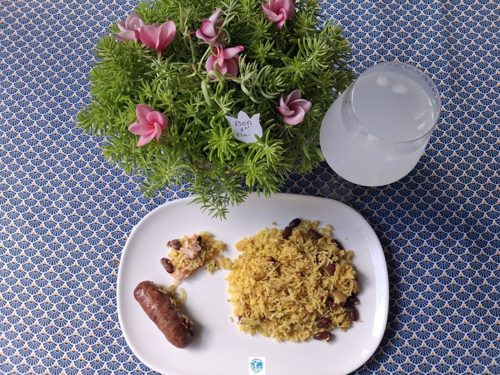 Assiette de riz jaune et saucisse frite sur table décorée de fleurs de frangipanier.