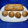 Gros plan en plongée d'une assiette ovale blanche remplie de pâtisseries maison dorées : un cake long aux pépites de chocolat et quatre petits gâteaux individuels, dont deux dômes retournés bombés et deux dômes lisses aux pépites. L'assiette est posée sur un fond bleu texturé.