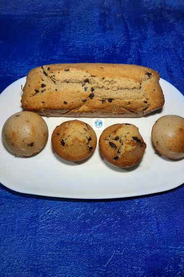 Gros plan en plongée d'une assiette ovale blanche remplie de pâtisseries maison dorées : un cake long aux pépites de chocolat et quatre petits gâteaux individuels, dont deux dômes retournés bombés et deux dômes lisses aux pépites. L'assiette est posée sur un fond bleu texturé.