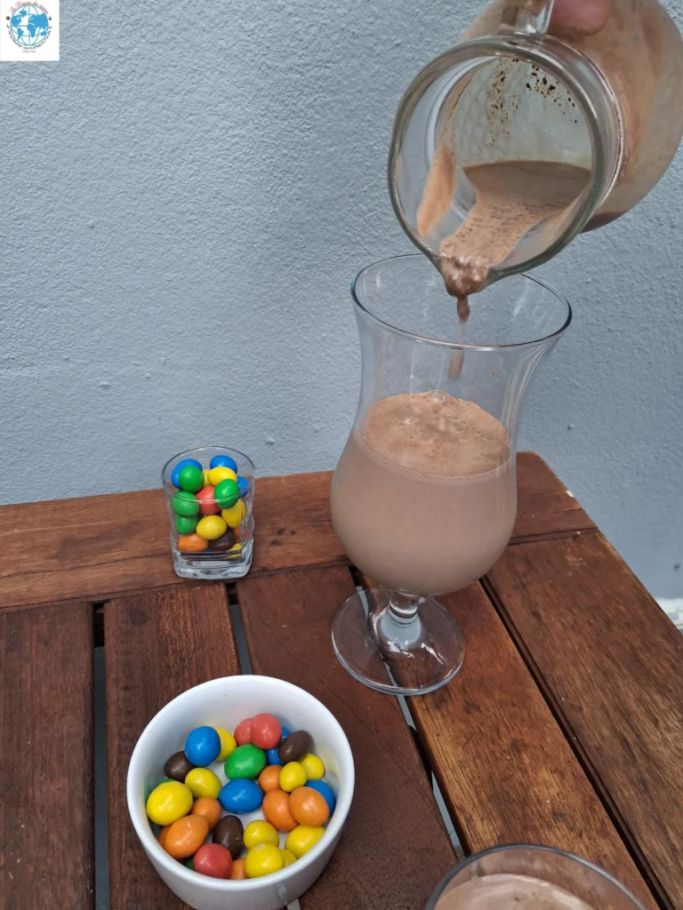 Un filet de lait au chocolat onctueux coule d'un pichet en verre dans un verre à pied sur une table en bois, accompagné d'un bol blanc et d'un petit verre remplis de M&M's colorés.