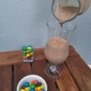 Un filet de lait au chocolat onctueux coule d'un pichet en verre dans un verre à pied sur une table en bois, accompagné d'un bol blanc et d'un petit verre remplis de M&M's colorés.