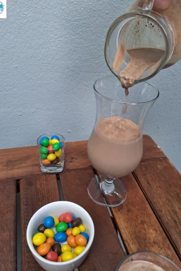 Un filet de lait au chocolat onctueux coule d'un pichet en verre dans un verre à pied sur une table en bois, accompagné d'un bol blanc et d'un petit verre remplis de M&M's colorés.