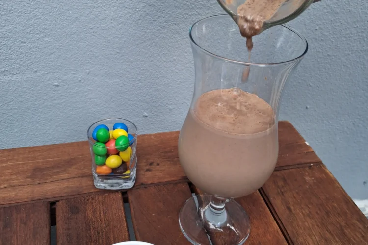 Un filet de lait au chocolat onctueux coule d'un pichet en verre dans un verre à pied sur une table en bois, accompagné d'un bol blanc et d'un petit verre remplis de M&M's colorés.