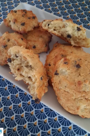 Une vue en plongée montre un plateau rectangulaire blanc garni de plusieurs biscuits dorés aux pépites de chocolat. Certains biscuits sont présentés entiers, tandis que d'autres sont disposés en deux morceaux, révélant une texture intérieure moelleuse et aérée. L'ensemble est posé sur une nappe ornée d'un motif géométrique répétitif en forme d'éventails bleu foncé et jaune sur fond bleu clair.