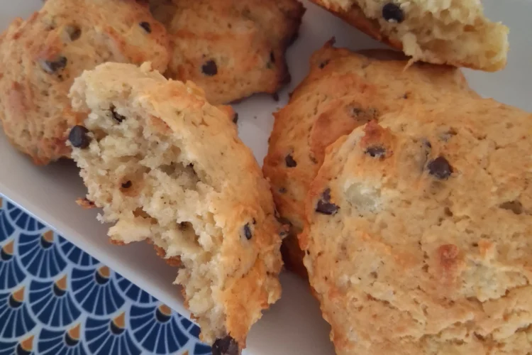 Une vue en plongée montre un plateau rectangulaire blanc garni de plusieurs biscuits dorés aux pépites de chocolat. Certains biscuits sont présentés entiers, tandis que d'autres sont disposés en deux morceaux, révélant une texture intérieure moelleuse et aérée. L'ensemble est posé sur une nappe ornée d'un motif géométrique répétitif en forme d'éventails bleu foncé et jaune sur fond bleu clair.